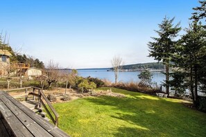 Eine grüne Holzterrasse mit Geländer, die auf einen See mit umliegenden Bäumen und einem Gebäude blickt.