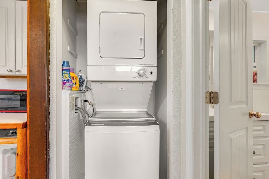 Un lavadero con lavadora y secadora apiladas, un estante con productos de limpieza y una puerta que da a otra habitación.