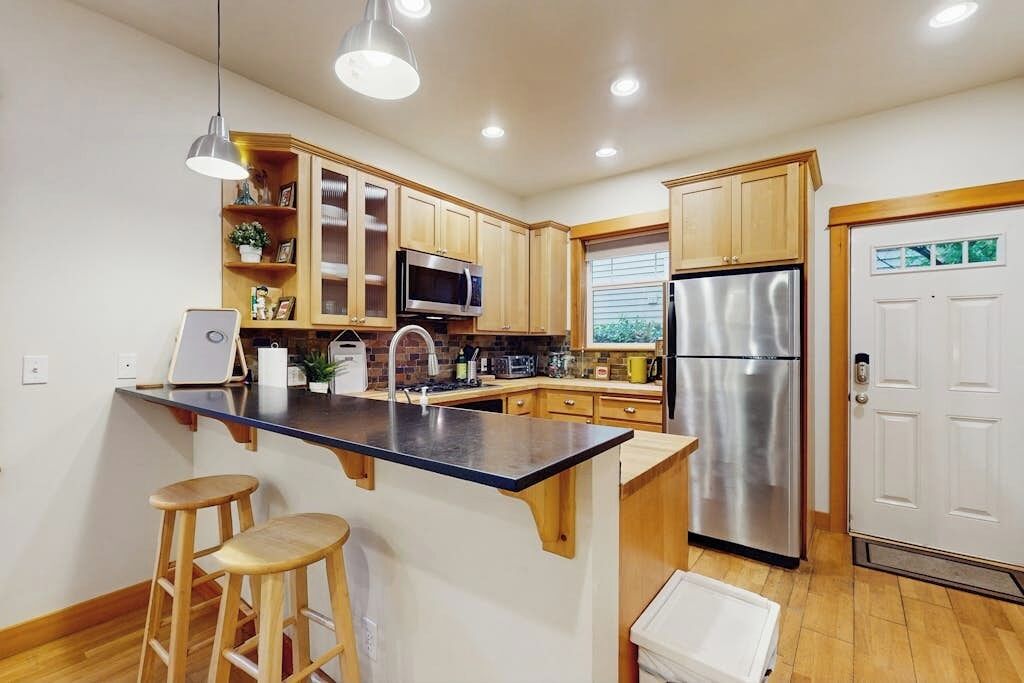 Una cocina con armarios de madera, un refrigerador de acero inoxidable, un microondas y un lavamanos. Hay dos taburetes y un balde blanco sobre la encimera.