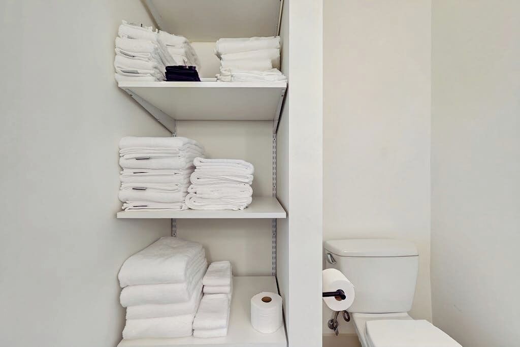 A bathroom with white shelves holding folded towels, a toilet, and a roll of toilet paper.