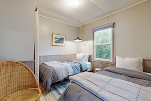 Un dormitorio con dos camas, una silla de mimbre, un ventanal con vistas y un cuadro en la pared.
