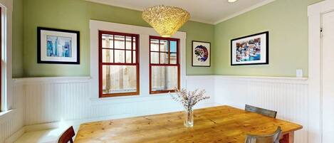 A dining room with a wooden table, white paneled walls, and two windows with red frames.
