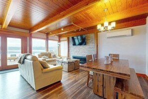 A spacious living area with a wooden ceiling, a large TV mounted on a brick wall, and a dining table with chairs.