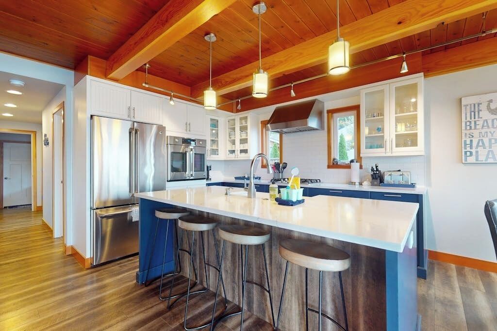 A modern kitchen with a central island, bar stools, stainless steel appliances, and wooden ceiling beams.