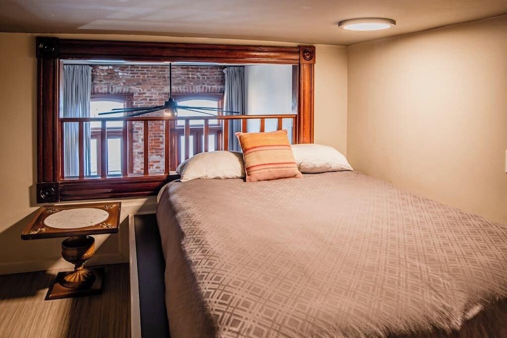 A hotel room with a large bed, a wooden headboard, a window with a view of a brick building, and a small table with a lamp.