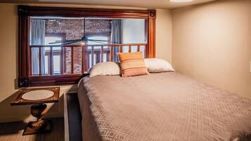 A hotel room with a large bed, a wooden headboard, a window with a view of a brick building, and a small table with a lamp.