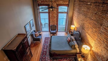 A bedroom with a large bed, a desk, a chair, a window with a view, and a brick wall.