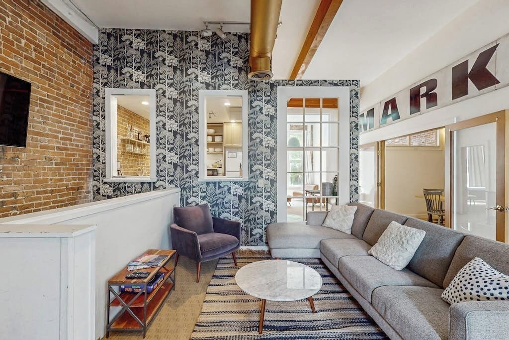 Un salon moderne avec un mur en briques, un papier peint à motifs, un canapé gris, une table basse ronde et une bibliothèque.
