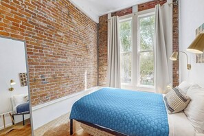 Una camera da letto con parete in mattoni, un'ampia finestra con tende e un letto con copriletto blu, oltre a una lampada da comodino.
