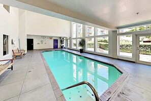 Ein Swimmingpool mit klarem, blauem Wasser, umgeben von einer gefliesten Terrasse, großen Fenstern und einigen Kübelpflanzen.