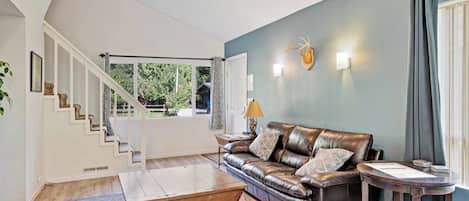 A modern living room with a leather sofa, a coffee table, a staircase, and a view of greenery outside.