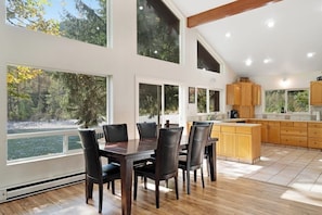 A modern kitchen with large windows, a dining area with a table and chairs, and wooden cabinets.