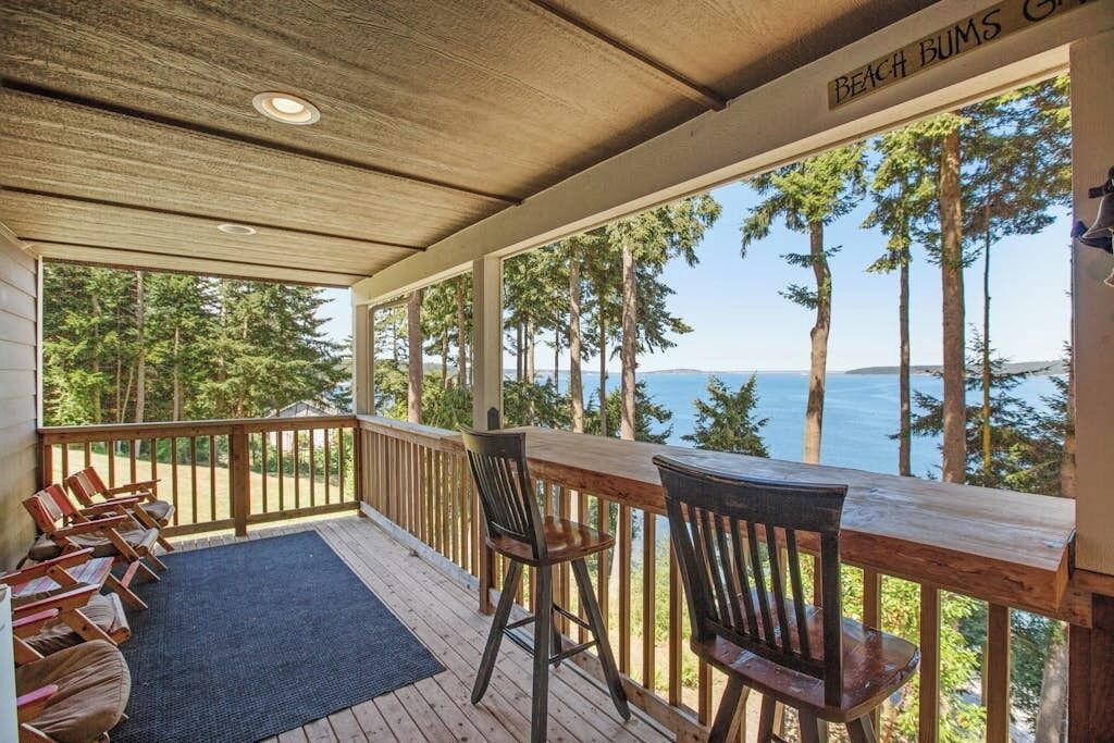 Une terrasse couverte, dotée de chaises et d’une table en bois, qui offre une vue sur une forêt et un plan d’eau.