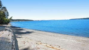 A beach with clear blue water, a wooden log, and a distant shoreline. - 3BR Bayview | Hot Tub | Deck | Firepit (Port Hadlock)