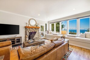 A living room with a large flat-screen TV, a stone fireplace, a leather sofa, and a view of the ocean through large windows. - Stunning 5BR Oceanview Birch Bay Dog Friendly (Blaine)
