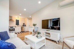A modern living room with a sofa, coffee table, and a TV. There is a kitchen in the background with white cabinets and a stove. - 1BR All-New - Central AC - Full Kitchen (Seattle)