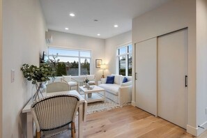 A modern living room with a white sofa, a dining table with chairs, and a large window providing a view of the outdoors. - 1BR All-New - Central AC - Full Kitchen (Seattle)