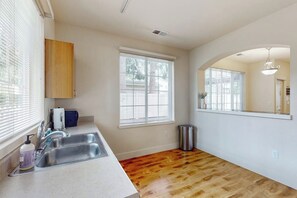 A kitchen with a wooden floor, a sink, a trash can, and a window with blinds. - Bright 2BR with deck, office & central location (Redmond)