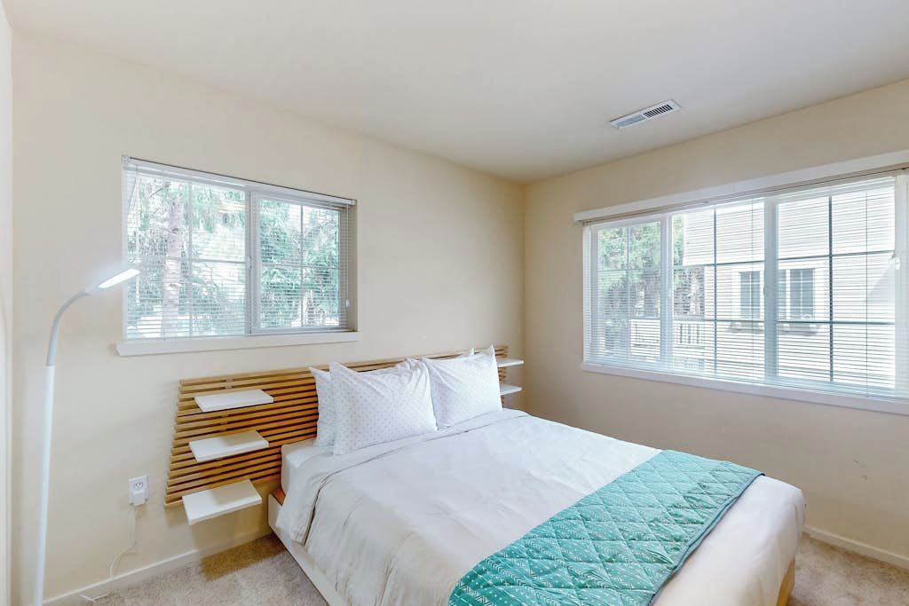 Un dormitorio con cama, dos ventanas, una lámpara de noche y estante en la pared.