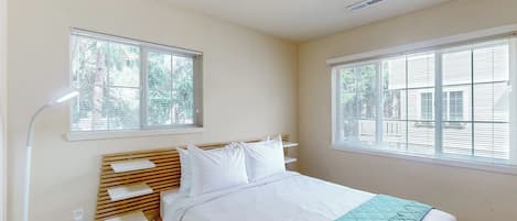 Un dormitorio con cama, dos ventanas, una lámpara de noche y estante en la pared.
