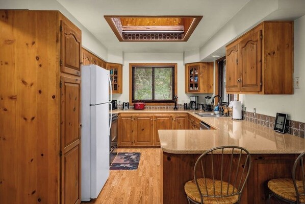 Une cuisine avec des meubles de rangement en bois, un réfrigérateur blanc, une fenêtre avec des stores et un comptoir avec des chaises.