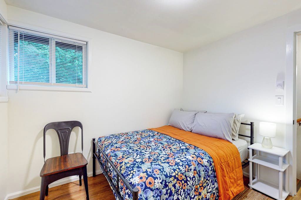 A bedroom with a bed, a chair, a bedside table with a lamp, and a window with blinds.