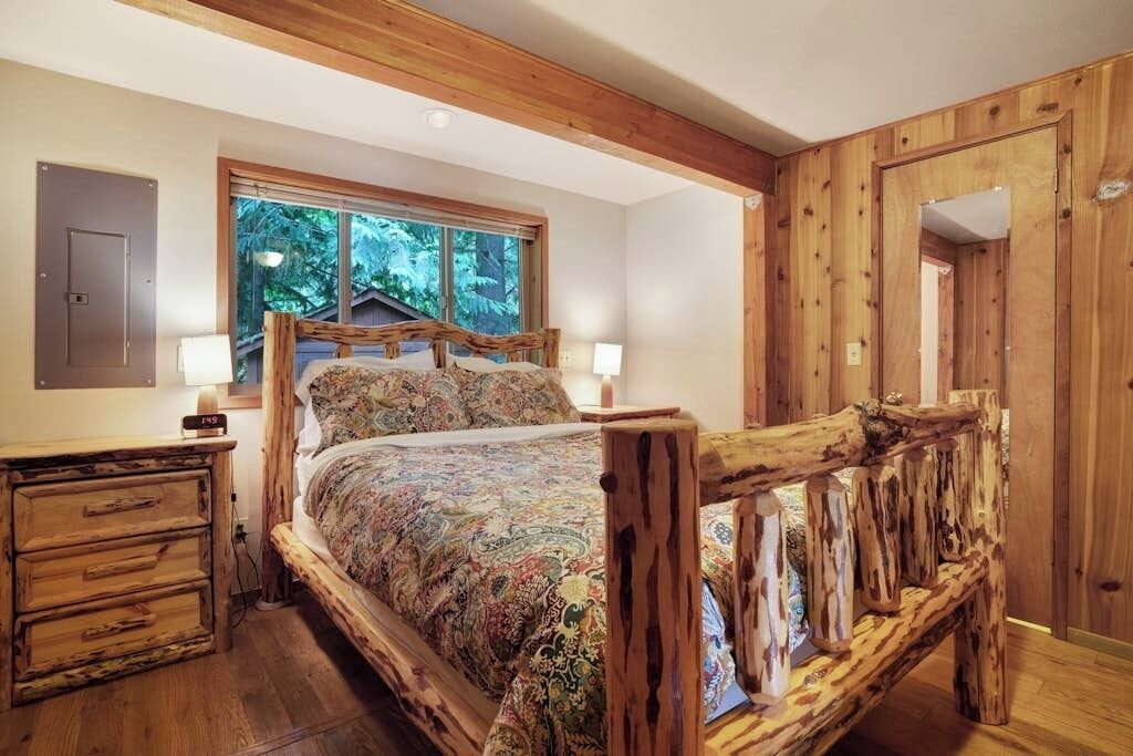 A bedroom with a wooden bed frame, a nightstand, and a window with a view of trees.