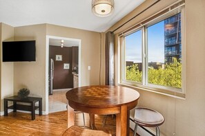 Ein Zimmer mit einem Holztisch, einem Stuhl, einem an der Wand montierten Fernseher und einem Fenster mit Blick auf Grünflächen und Gebäude.