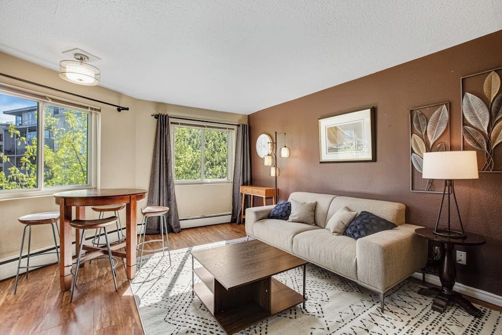 A modern living room with a sofa, coffee table, and bar stools. There is a large window with curtains, a lamp, and a framed picture on the wall.