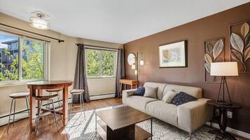 A modern living room with a sofa, coffee table, and bar stools. There is a large window with curtains, a lamp, and a framed picture on the wall.