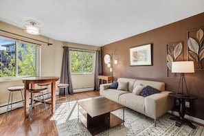 Ein modernes Wohnzimmer mit einer Couch, einem Couchtisch und Barhocker. Es gibt ein großes Fenster mit Vorhängen, eine Lampe und ein gerahmtes Bild an der Wand.