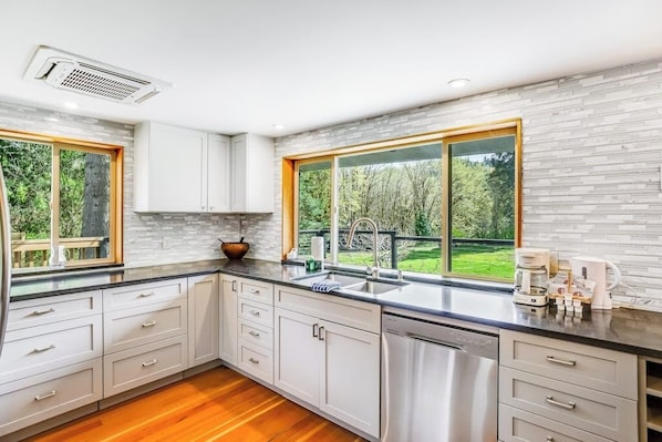 A modern kitchen with white cabinetry, a dishwasher, and a large window offering a view of greenery.