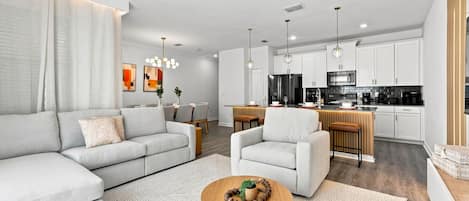 A modern living room with a sofa, armchair, and coffee table, a dining area with chairs, and a kitchen with appliances.