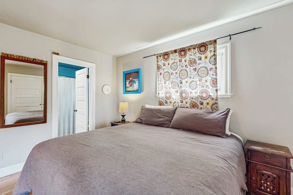 A bedroom with a bed, a nightstand, a lamp, a window with patterned curtains, a door, and a wall clock.