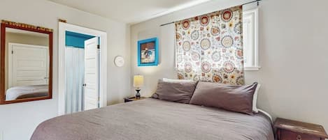 A bedroom with a bed, a nightstand, a lamp, a window with patterned curtains, a door, and a wall clock.