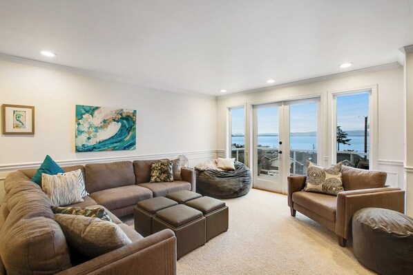 A living room with a sectional sofa, a bean bag, and a view of the ocean through large windows. - 5BR Oceanview - Hot Tub - Fireplace - Balcony (Camano Island)