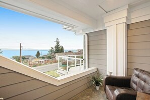 A room with a view of a residential street and a body of water, a window with a white frame, a brown leather sofa, and a small potted plant. - 5BR Oceanview - Hot Tub - Fireplace - Balcony (Camano Island)