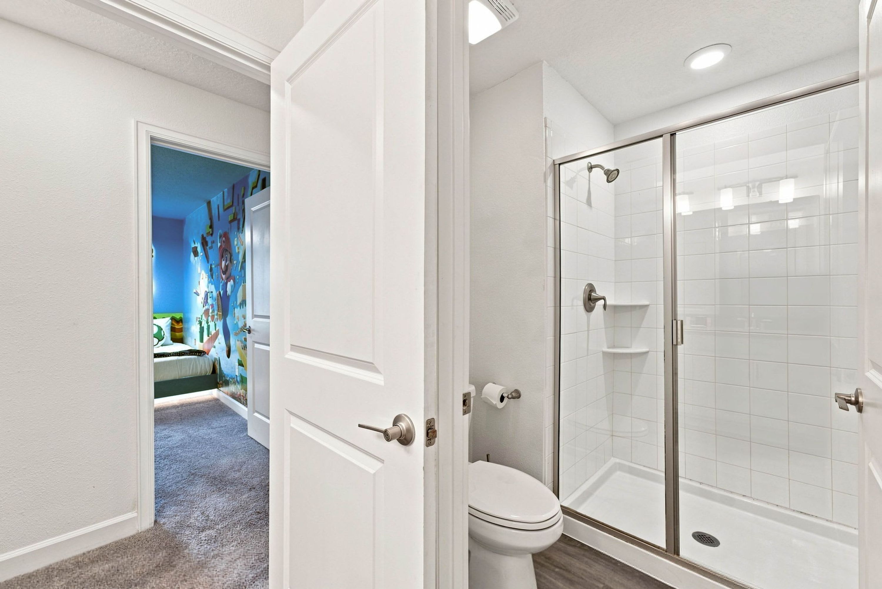 Un bagno con il water, una doccia con ante in vetro e un corridoio che porta a una camera da letto con un murale colorato.