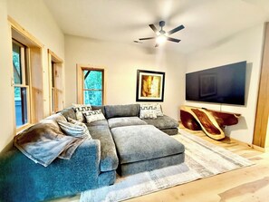 A modern living room with a sectional sofa, a ceiling fan, a flat-screen TV, and a large window. - Fantastic 4-bedroom, Spacious Timberframe Cabin in charming Lake Lure (Lake Lure)