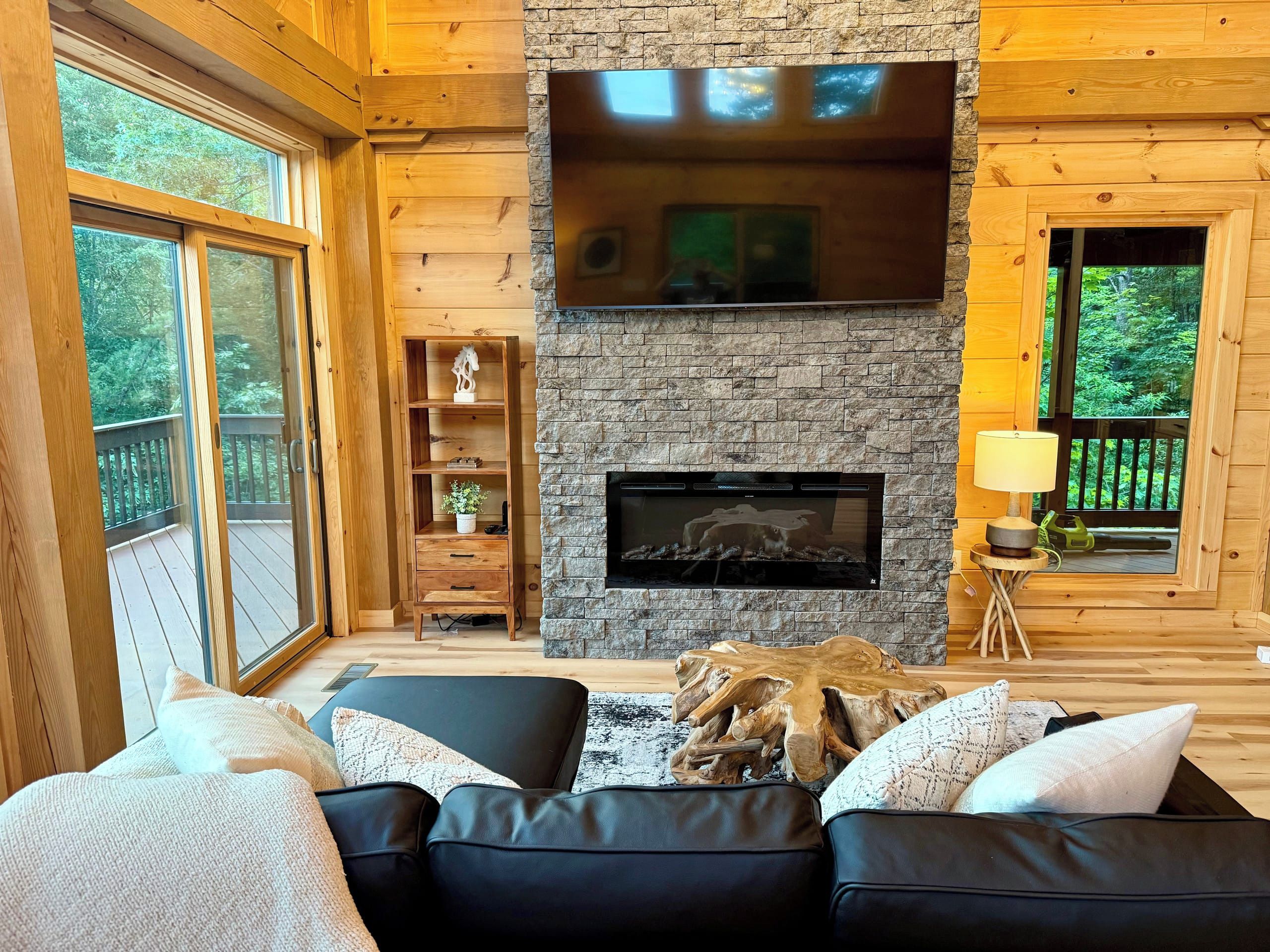 A cozy living room with a fireplace, wooden walls, and a large window overlooking greenery.