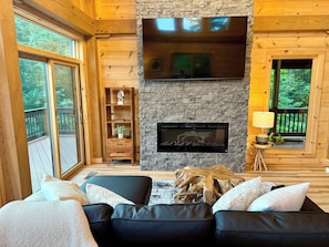 A cozy living room with a fireplace, wooden walls, and a large window overlooking greenery. - Fantastic 4-bedroom, Spacious Timberframe Cabin in charming Lake Lure (Lake Lure)