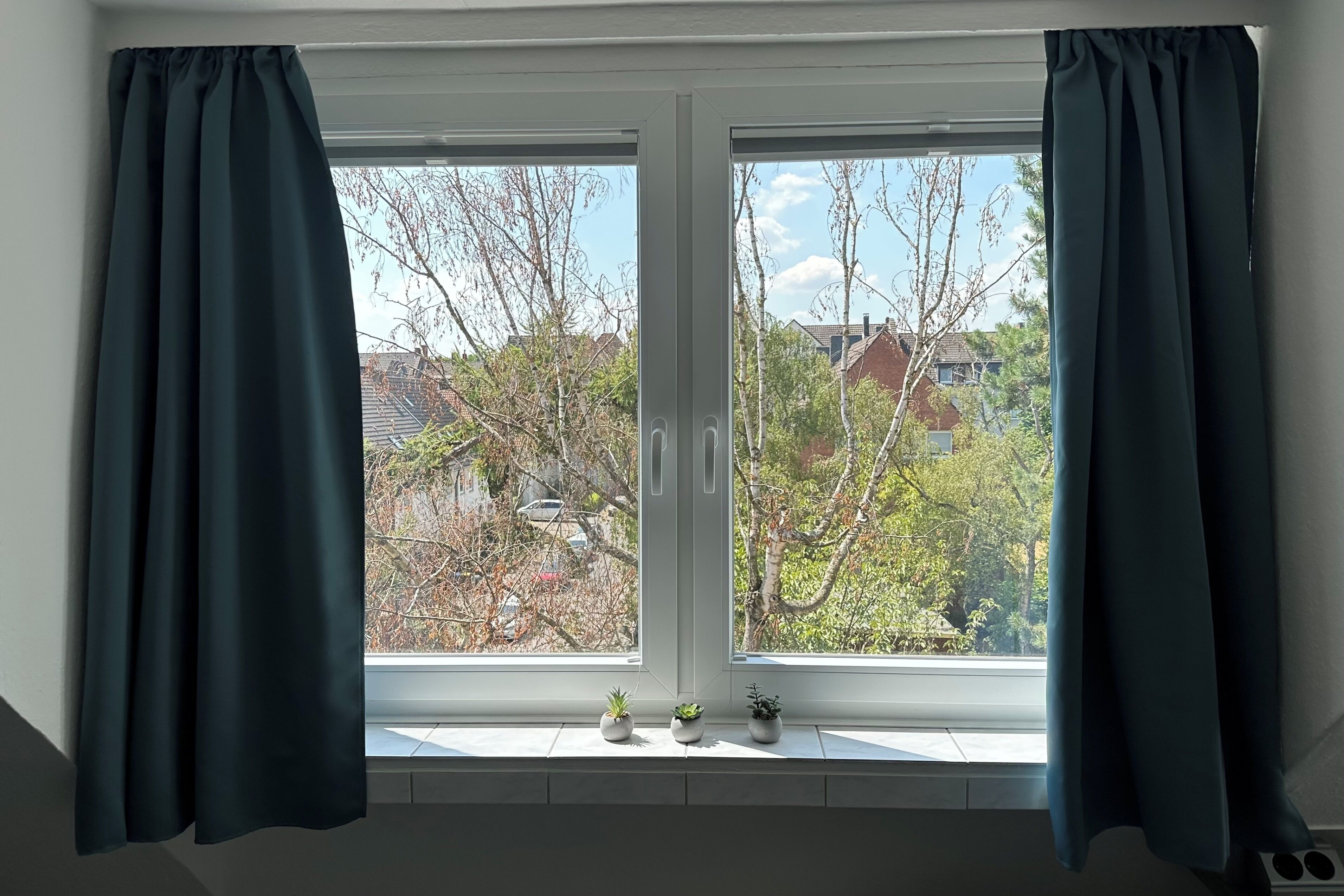 A window with two curtains, a view of trees and houses, and three small potted plants on the windowsill.