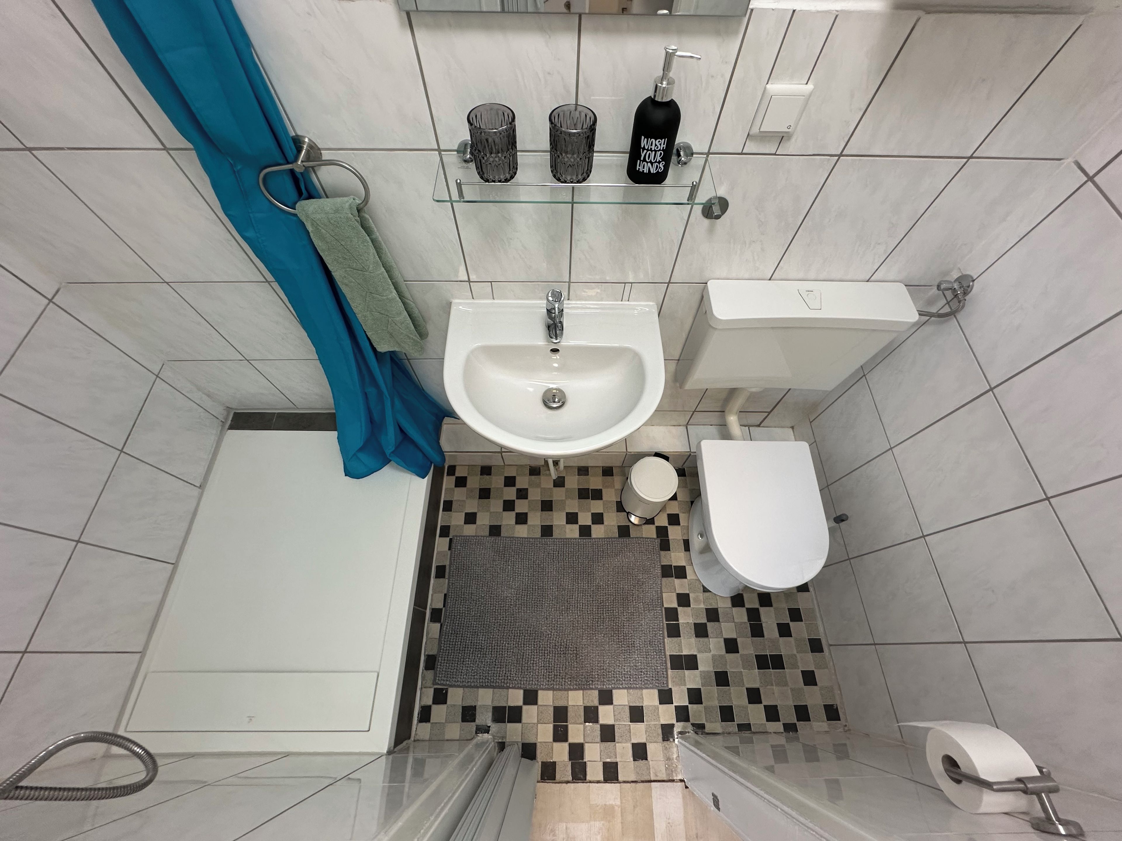 Ein Badezimmer mit Waschbecken, Toilette und Dusche. Ein Handtuchhalter, eine Ablage mit Toilettenartikeln und ein Schachbrettmuster-Boden sind vorhanden.