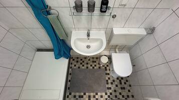 Ein Badezimmer mit Waschbecken, Toilette und Dusche. Ein Handtuchhalter, eine Ablage mit Toilettenartikeln und ein Schachbrettmuster-Boden sind vorhanden.