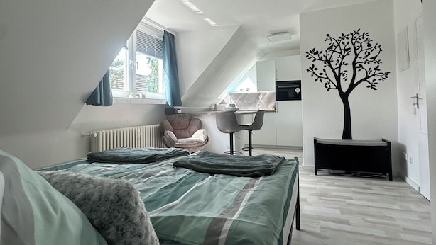 Une chambre à coucher comprenant un lit, une chaise, un radiateur, une fenêtre avec des rideaux et un motif décoratif représentant un arbre sur un mur.