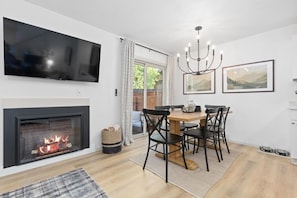 A modern living room with a fireplace, a dining table with chairs, a chandelier, and a flat-screen TV mounted on the wall.