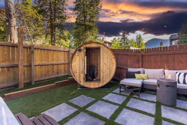 A wooden sauna in a backyard with a wooden fence, a stone patio, and a sofa with cushions.