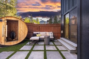 A wooden sauna with a glass door, a seating area with cushions, a fire pit, and a stone pathway.