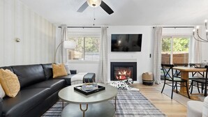 Ein Wohnzimmer mit einem schwarzen Ledersofa, einem runden Couchtisch, einem Kamin, einem Esstisch mit Stühlen und einem an der Wand montierten Fernseher.