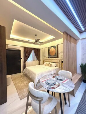 A modern bedroom with a bed, bedside tables, a round dining table with chairs, a ceiling fan, and an air conditioning unit. - Estaaron Crave - Villas and Apartment Hotel (Tema)